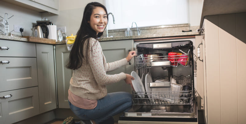 Picture of woman crouching down by her open dishwasher, smiling to camera.