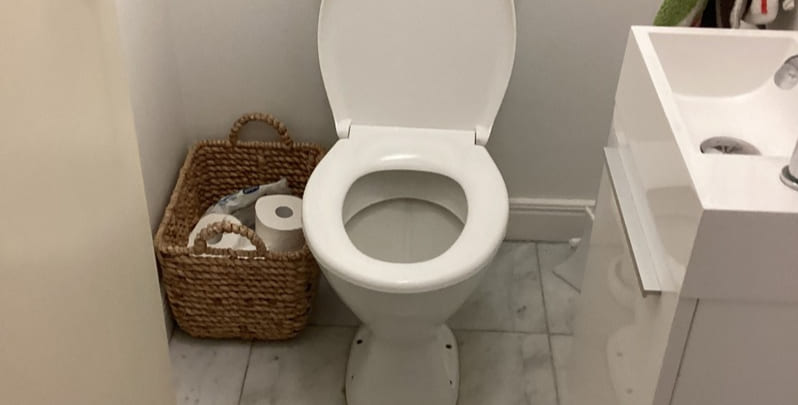 Unblocked toilet with lid up next to it is a wooden basket filled with toilet paper rolls.